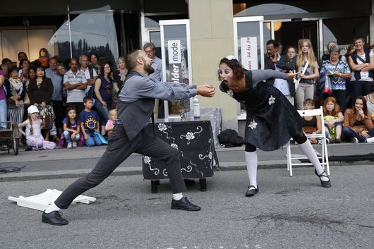 Zwei Strassenkünstler in schwarz-grauer Kostümierung führen eine dramatische Theaterszene auf einem belebten Stadtplatz auf. Eine Frau in schwarzem Kleid beugt sich weit vor, während ein Mann in grauem Anzug ihr entgegenlehnt, umgeben von einem grossen Publikum.