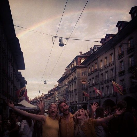 Drei junge Menschen jubeln mit ausgestreckten Armen auf einer belebten Stadtstrasse, während ein Regenbogen über historischen Gebäuden am bewölkten Himmel erscheint.