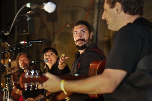 Eine Musikgruppe spielt gemeinsam auf einer kleinen Bühne: Ein bärtiger Mann mit Gitarre blickt lächelnd zu einem Mitspieler, während eine Frau in Rot ins Mikrofon singt.