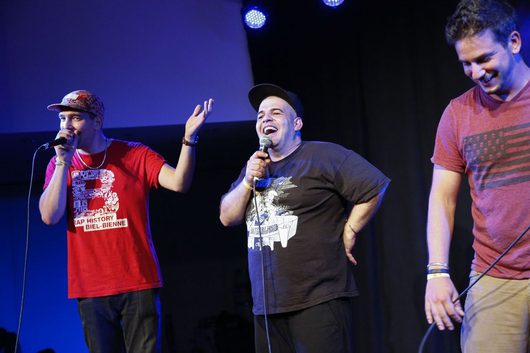 Drei Männer stehen auf einer Bühne und performen gemeinsam, zwei von ihnen halten Mikrofone und lachen ausgelassen. Der mittlere Performer trägt eine schwarze Kappe und ein schwarzes T-Shirt und gestikuliert enthusiastisch.