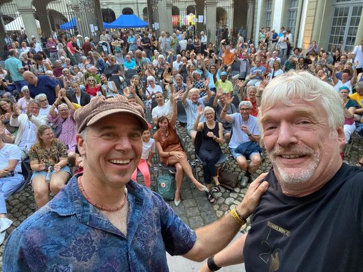 Zwei lächelnde Männer machen ein Selfie vor einer grossen, winkenden Menschenmenge auf einem Innenhof-Konzert. Im Hintergrund sind Hunderte von Zuschauern auf Stühlen zu sehen, die fröhlich in die Kamera winken.