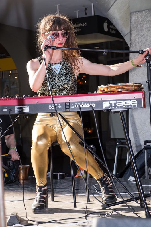 Eine Musikerin mit goldenen Glanzleggings und rosa Sonnenbrille singt in ein Mikrofon und spielt gleichzeitig ein rotes Stage-Eighty-Eight-Keyboard bei einem Outdoor-Auftritt.
