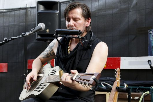 Ein tätowierter Musiker spielt eine Resonatorgitarre und bläst gleichzeitig Mundharmonika über einen Mundharmonikahalter vor einem Mikrofon auf einer Freiluftbühne. Er trägt ein ärmelloses schwarzes Shirt und eine bunte Perlenkette.