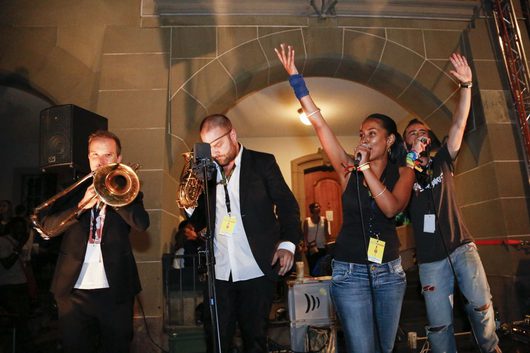 Eine Band tritt bei einem Abendkonzert in einem historischen Gebäude auf: Ein Musiker spielt Posaune, ein weiterer beugt sich zum Mikrofon, während eine Sängerin und ein Rapper mit erhobenen Händen performen.