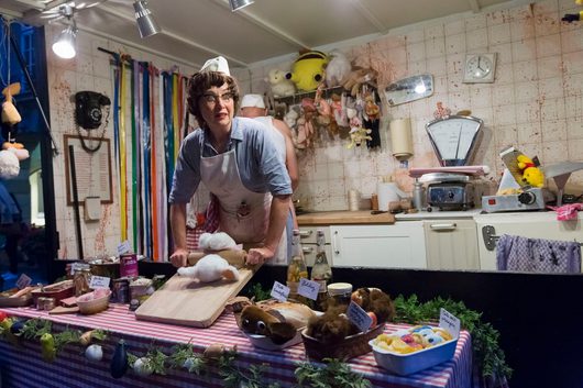 Eine als Metzgerin verkleidete Frau mit weisser Schürze und Haube steht hinter einem mit Speisen und Requisiten dekorierten Tisch in einer surrealen, theatralischen Metzgerei-Kulisse. Die Wände sind mit roten Flecken besprenkelt und mit Stofftieren behängt, was der Szene einen absurden, bühnenhaften Charakter verleiht.