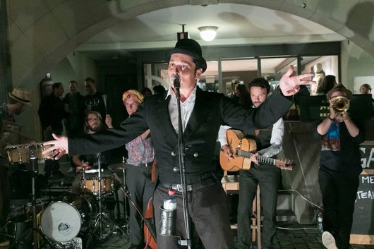 Ein Sänger mit schwarzem Hut und Weste steht mit ausgebreiteten Armen vor einem Mikrofon und performt mit seiner Band in einem gewölbten Durchgang. Im Hintergrund sind ein Gitarrist, ein Trompeter, ein Schlagzeuger und mehrere Zuschauer zu sehen.