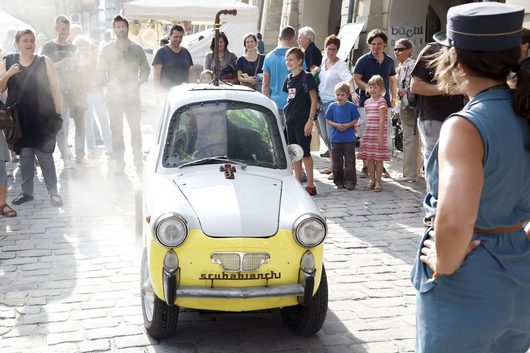 Ein kleines, weiss-gelbes Oldtimer-Fahrzeug mit der Aufschrift «scubabianchi» fährt auf einem Kopfsteinpflaster durch eine belebte Fussgängerzone, umgeben von neugierigen Zuschauern. Dampf oder Rauch steigt vom Fahrzeug auf, während Kinder und Erwachsene das ungewöhnliche Spektakel beobachten.