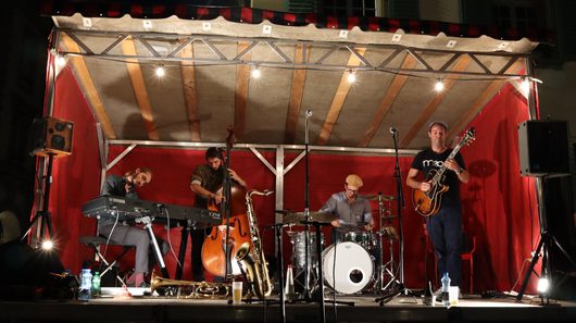 Ein Jazz-Quartett tritt auf einer kleinen, rot dekorierten Freiluftbühne auf, mit Keyboard, Kontrabass, Schlagzeug und E-Gitarre. Auf dem Bühnenboden liegen zusätzliche Instrumente wie Saxofon und Trompete.