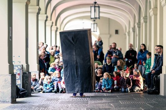 Eine Person versteckt sich hinter einer grossen schwarzen Tafel und tritt in einem historischen Laubengang vor einem Publikum aus Kindern und Erwachsenen auf. Die Zuschauer sitzen und stehen entlang der Arkaden und beobachten die Strassentheatervorstellung aufmerksam.