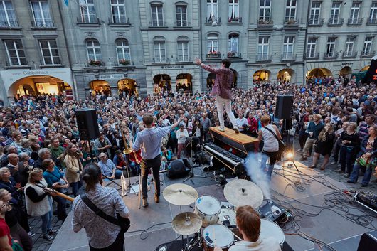 Eine Band spielt auf einer Freiluftbühne vor einer riesigen Menschenmenge auf einem historischen Stadtplatz. Ein Sänger steht auf einem Klavier und streckt die Arme aus, während Hunderte von Zuschauern zuschauen.