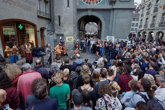 Eine Strassenband namens 'Shishko Disco' spielt vor dem Zytglogge in Bern und wird von einer grossen Menschenmenge umringt. Die Musiker tragen auffällige, bunte Kostüme und spielen verschiedene Instrumente auf dem Kopfsteinpflaster.