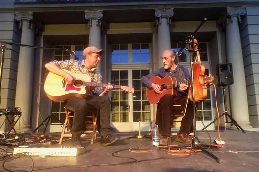 Zwei Männer sitzen auf einer Bühne vor einem klassizistischen Gebäude und spielen akustische Gitarren bei einem Abendkonzert im Freien. Der linke Musiker trägt eine Kappe und ein gemustertes Hemd, der rechte hat langes graues Haar und einen Bart.