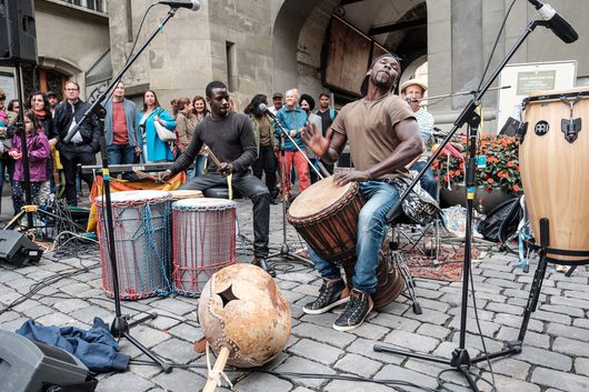 Zwei Musiker spielen afrikanische Trommeln bei einem Strassenkonzert auf einem Kopfsteinpflasterplatz in einer europäischen Altstadt, umgeben von einer aufmerksamen Zuschauermenge.