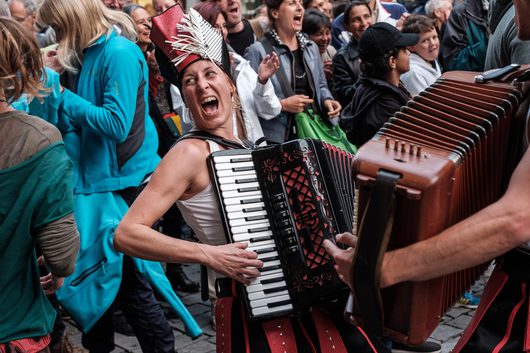 Eine Frau mit einem auffälligen roten Federhut spielt leidenschaftlich Akkordeon auf einer belebten Strassenveranstaltung. Sie singt mit weit geöffnetem Mund, umgeben von einer begeisterten Menschenmenge.