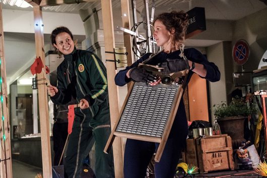 Zwei Frauen agieren in einer Theaterperformance inmitten eines selbstgebauten Holzgestells mit verschiedenen Alltagsgegenständen und Werkzeugen. Die linke Frau lächelt breit in einem grünen Overall, während die rechte eine Waschbrettschürze und Küchenutensilien trägt.