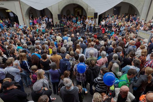 Eine grosse Menschenmenge versammelt sich auf einem Stadtplatz in Bern, um eine Band beim Buskers Bern Strassenmusikerfestival zuzuhören. Die Musiker spielen auf einer improvisierten Bühne unter historischen Laubenbögen.