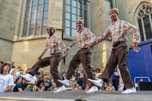Drei Männer in identischen karierten Hemden und braunen Hosen tanzen synchron auf einer Freilichtbühne vor einer gotischen Kirche. Im Hintergrund verfolgt eine grosse Menschenmenge aus Kindern und Erwachsenen die energetische Darbietung.