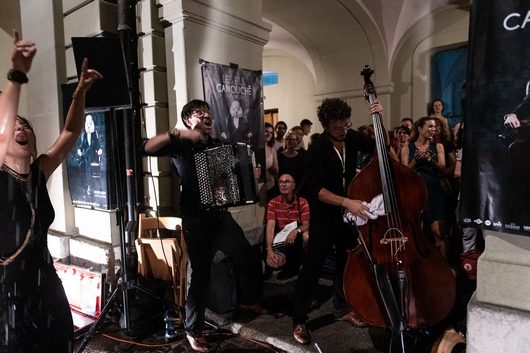 Ein Akkordeonist und ein Kontrabassist spielen leidenschaftlich bei einem Livekonzert unter einem Bogendurchgang, während eine Frau links mit erhobenen Armen mitfeiert und das Publikum im Hintergrund begeistert reagiert.