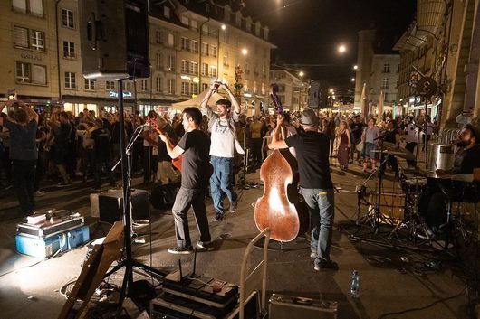 Eine Band spielt nachts auf einer belebten Stadtstrasse vor einer grossen Menschenmenge, mit einem Kontrabass, Geige und Schlagzeug im Vordergrund. Die Musiker bewegen sich energetisch auf dem Pflaster, während das Publikum im Hintergrund tanzt und feiert.