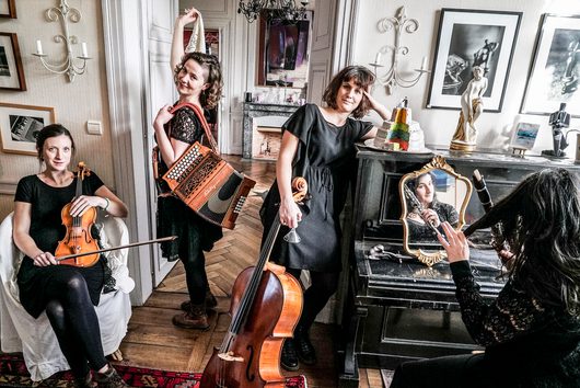 Vier Musikerinnen in schwarzer Kleidung posieren in einem eleganten Salon mit Geige, Akkordeon, Cello und Klarinette. Eine der Musikerinnen ist im Spiegel eines schwarzen Klaviers reflektiert.