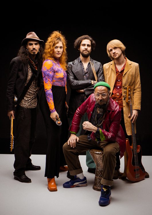 Fünf Musikerinnen und Musiker posieren gemeinsam vor einem schwarzen Studiohintergrund, gekleidet in farbenfrohe, eklektische Outfits. Zwei von ihnen halten Gitarren, während ein älterer Mann in der Mitte auf einem verzierten Hocker sitzt.