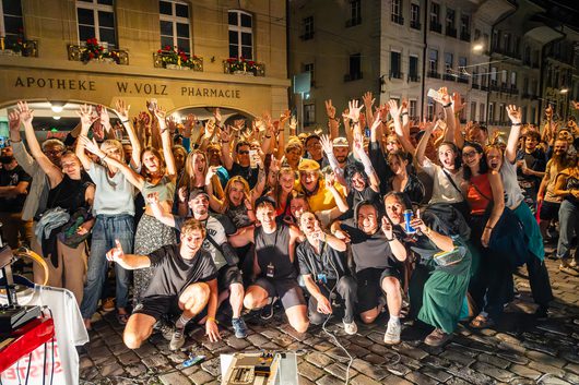 Eine grosse, ausgelassene Menschenmenge posiert nachts auf einem Kopfsteinpflasterplatz vor einer Apotheke und streckt jubelnd die Arme in die Höhe. Im Vordergrund sind Musikequipment und Effektpedale auf dem Boden zu sehen.