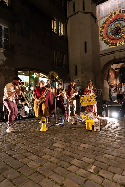 Eine Brassband namens «Perhaps Contraption» spielt nachts auf einem Kopfsteinpflaster-Platz in einer Altstadt, im Hintergrund ist eine historische astronomische Uhr zu sehen. Die Musizierenden tragen farbenfrohe Kostüme und spielen Saxophone, Posaune und Tuba.
