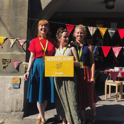 Drei lächelnde Frauen stehen nebeneinander vor einem Gebäude und halten ein gelbes Schild mit der Aufschrift «Buskers Bern – Vesselil, Nordic Vocal String Folk (DK)». Im Hintergrund sind bunte Wimpelketten und ein Marktstand zu sehen.