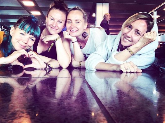 Vier junge Frauen lehnen lächelnd auf einer glänzenden Tischfläche und posieren gemeinsam für ein Gruppenfoto. Eine von ihnen formt mit ihren Händen ein Herz.