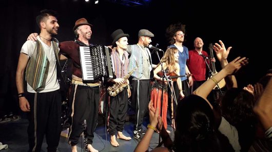 Eine Musikband mit sieben Mitgliedern steht nach einem Konzert auf der Bühne und verbeugt sich vor dem jubelnden Publikum. Die Musiker tragen bunte, folkloristische Kostüme und halten Instrumente wie Akkordeon, Saxofon, Geige und Gitarre.
