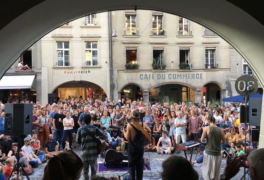 Eine grosse Menschenmenge verfolgt ein Strassenkonzert auf einem Stadtplatz, gerahmt durch einen Rundbogen. Zwei Musikerinnen oder Musiker mit Gitarren stehen vor dem Publikum, im Hintergrund sind das Café du Commerce und das Geschäft Kunstreich zu sehen.