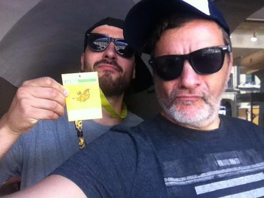 Zwei Männer mit Sonnenbrillen und Caps machen ein Selfie, wobei einer von ihnen ein gelbes Ticket oder Abzeichen in die Kamera hält.