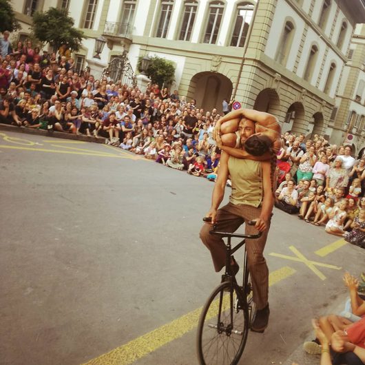 Ein männlicher Akrobat fährt auf einem Fahrrad durch eine gesperrte Strasse und trägt dabei eine zweite Person auf seinen Schultern, während eine grosse Menschenmenge das Strassentheater-Spektakel beobachtet.