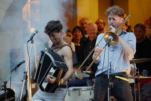 Zwei Musiker treten leidenschaftlich auf einer Bühne auf: links ein Mann mit freiem Oberkörper spielt Akkordeon, rechts ein Mann im Hemd bläst Posaune. Im Hintergrund verfolgt ein dicht gedrängtes Publikum die Darbietung.