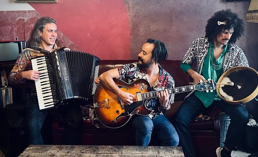 Drei Musiker spielen gemeinsam in einem rustikalen Innenraum: ein Mann mit Akkordeon, einer mit einer Archtop-Gitarre und einer mit einem Rahmentrommel-Instrument. Die Gruppe wirkt entspannt und lebendig.