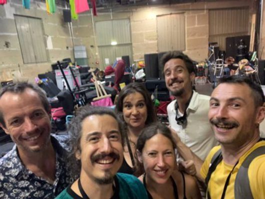 Sechs lächelnde Erwachsene machen ein Gruppen-Selfie in einer grossen Halle mit Musikequipment im Hintergrund. Die Gruppe wirkt fröhlich und entspannt in einer Veranstaltungs- oder Festivalatmosphäre.
