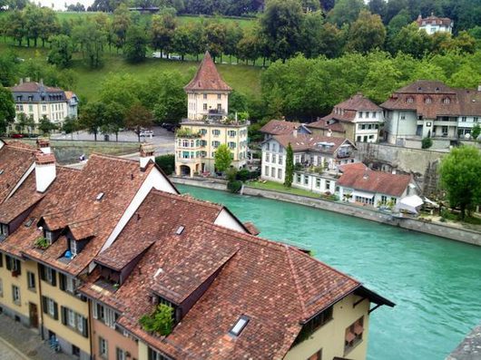 Luftaufnahme der Altstadt von Bern mit türkisfarbenem Flusslauf der Aare, roten Ziegeldächern historischer Gebäude und üppig begrünten Hügeln im Hintergrund.
