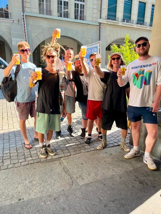 Eine Gruppe von sieben jungen Männern stösst mit Plastikbechern Bier auf einem gepflasterten Stadtplatz an. Sie lachen und heben ihre Becher in die Kamera, im Hintergrund ist ein klassisches Stadtgebäude zu sehen.