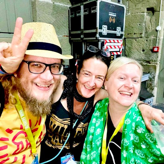 Drei lachende Personen machen ein Selfie hinter der Bühne eines Festivals, alle tragen gelbe Akkreditierungsbänder. Der Mann links trägt einen Strohhut und zeigt das Rock-Handzeichen.
