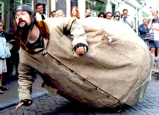 Ein Strassenkünstler mit Helm und Bart rollt einen riesigen, eiförmigen Gegenstand aus beigem Stoff über ein Kopfsteinpflaster, während eine lachende Menschenmenge zuschaut.