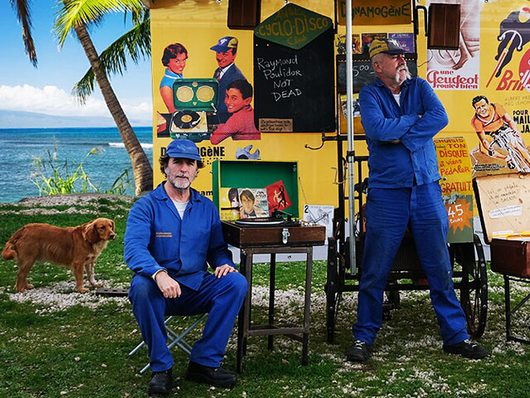 Zwei Männer in blauen Overalls posieren vor einem bunten, mit Vintage-Plakaten dekorierten Marktstand am Meer. Ein brauner Hund steht links im Bild, im Hintergrund sind eine Palme und das türkisfarbene Meer zu sehen.
