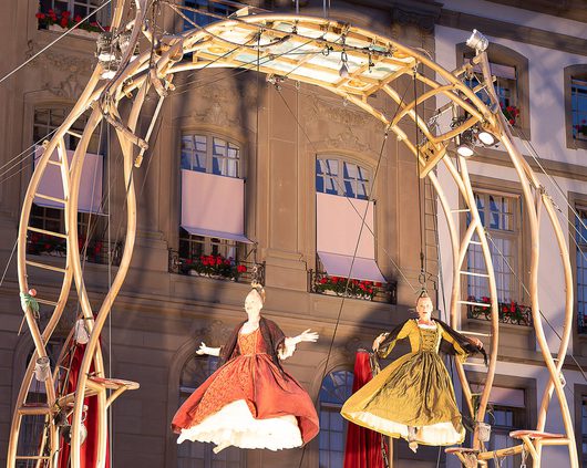 Zwei Darstellerinnen in historischen Barockkleidern schweben an Seilen in einem großen hölzernen Bogengestell vor einem klassizistischen Gebäude. Die Frauen tragen aufwendige Roben in Rot und Goldgrün und führen eine Freiluft-Theateraufführung auf.
