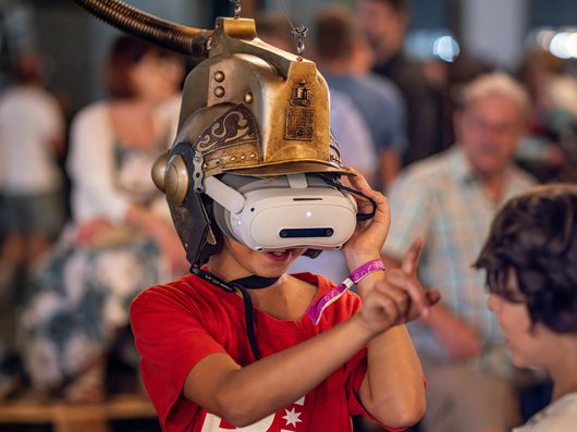 Ein Kind trägt eine aufwendige Steampunk-Helm-Konstruktion mit integrierter VR-Brille und hält die Hände vor sich, während es in eine virtuelle Realität eintaucht. Im Hintergrund sind unscharf mehrere Erwachsene und ein weiteres Kind zu sehen.
