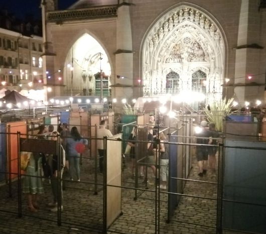Besucher schlendern abends durch einen Freiluftmarkt oder eine Ausstellung auf einem Kopfsteinpflasterplatz vor einer beleuchteten gotischen Kathedrale. Lichterketten und Scheinwerfer erhellen die Stände und Trennwände, während die Kirchenfassade mit ihren Spitzbögen im Hintergrund leuchtet.