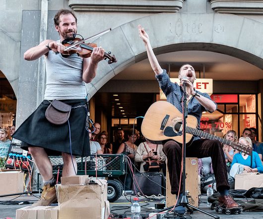 Zwei Strassenmusiker treten vor einem historischen Arkadenbogen auf: Ein Mann im Kilt spielt leidenschaftlich Geige, während sein Begleiter sitzend Gitarre spielt und mit ausgestrecktem Arm ins Mikrofon singt.