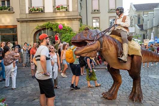 Ein Performer reitet auf einem lebensgrossen animatronischen Tyrannosaurus-Rex-Kostüm durch eine belebte Fussgängerzone in einer europäischen Altstadt, während Passanten staunen und Fotos machen.