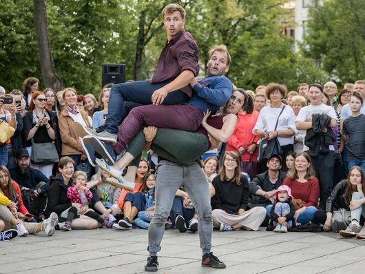 Drei Strassenartisten führen eine akrobatische Hebefigur vor einem grossen Publikum im Freien auf. Ein Mann trägt drei weitere Personen gleichzeitig auf einem gepflasterten Platz, umgeben von staunenden Zuschauern.