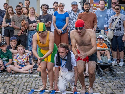 Zwei Personen in Schwimmkleidung und Bademützen beugen sich an einer Startlinie auf einem Kopfsteinpflasterplatz vor, während ein Mann mit Trillerpfeife als Starter kniet und eine grosse Menschenmenge zuschaut.