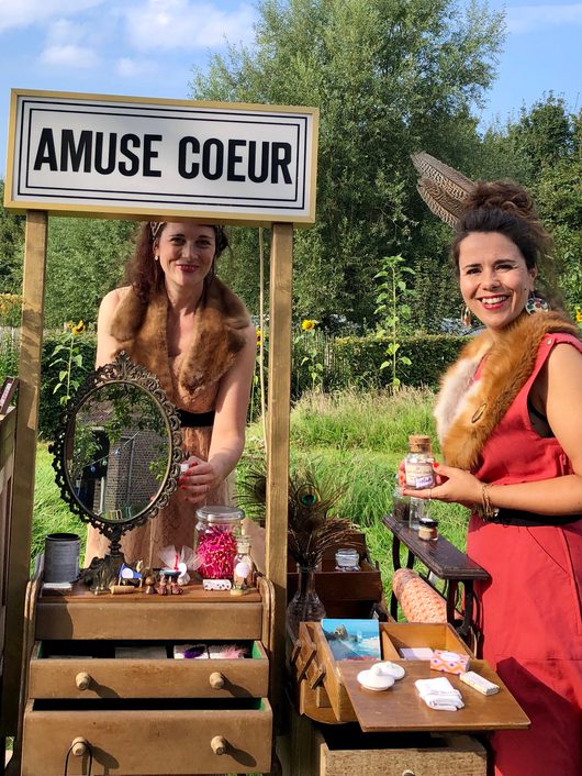 Zwei kostümierte Frauen stehen an einem dekorativen Marktstand mit der Aufschrift «Amuse Coeur» und präsentieren verschiedene kleine Objekte und Fläschchen. Der Stand ist mit einem antiken Spiegel, Gläsern und Schubladen ausgestattet und befindet sich im Freien vor einem grünen Garten.