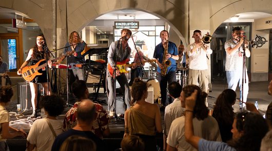 Eine siebenköpfige Band spielt ein Livekonzert unter einem Bogendurchgang in einer städtischen Passage, während ein dichtes Publikum zuschaut. Die Musiker spielen Bassgitarre, E-Gitarre, Keyboard, Saxofon und zwei Trompeten, begleitet von einem Sänger am Mikrofon.
